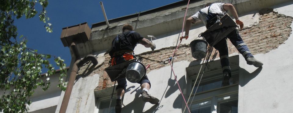 Dva výškoví pracovníci opravují omítku domu z lana